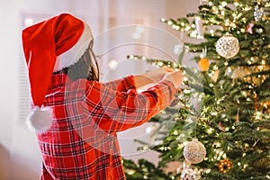Christmas tree Decorating. Girl child in santa hat and plaid pajamas decorate the Christmas tree with red shiny balls