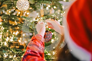 Christmas tree Decorating. Girl in santa hat and plaid pajamas decorate the Christmas tree with red shiny balls.hands