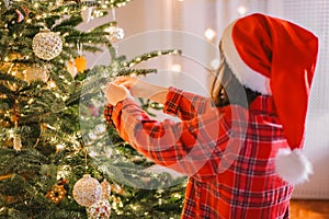 Christmas tree. Girl child in santa hat and plaid pajamas decorate the Christmas tree with red shiny balls.hands put