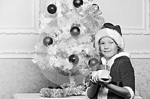 Christmas tree ideas for kids. Boy kid dressed as santa with red hat hold gift box near christmas tree. Christmas santa