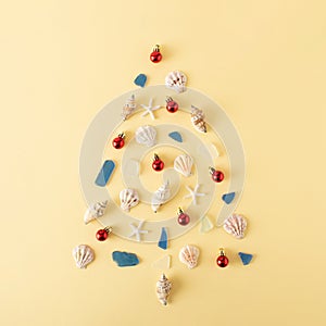 Christmas tree made of seashells, glass, starfish and red baubles on pastel yellow background. Creative minimal New Year tropics