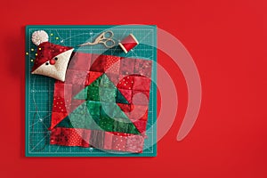 Christmas tree patchwork block, pincushion like Santa, scissors, spool of thread on craft mat, red background