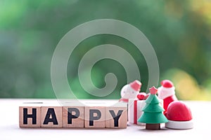 Christmas tree beside wooden block with `HAPPY` and nature background.