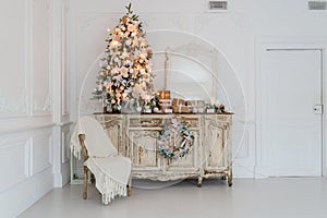 Christmas tree on wooden chest of drawers commode bureau in white interior, decorated with artificial flowers, garlands and toys