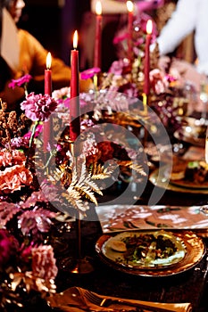 Christmas wedding dinner served table. Shiny leaves of gold paint, pink and red flowers and gold dishes. candles in a metal