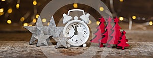 Christmas white alarm clock with vintage red wooden firtrees and brown grey stars with garland lights background, banner