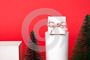 Christmas white gift box with tied bow stay on cube podium and decoration fir trees on red background