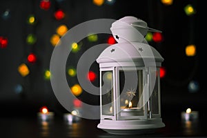 Christmas white lantern with a burning candle on the background of colorful lights garland, bokeh.