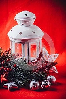 Christmas white lantern is standing with a burning candle in it with a fir tree branch and knit stars on a red background.