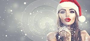 Beauty model woman in Santa`s hat blowing snow in her hand. Christmas winter fashion girl on holiday blurred winter background