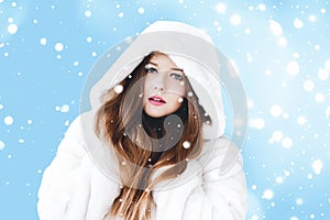 Christmas and winter holiday portrait of young woman in white hooded fur coat, snow on blue background, fashion and