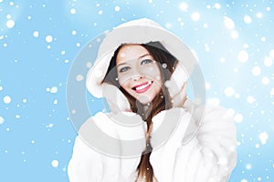 Christmas and winter holiday portrait of young woman in white hooded fur coat, snow on blue background, fashion and