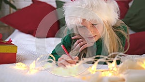 Christmas wish list. Smiling girl in Santa Claus hat writing letter dreams for gifts to Santa Claus. Child lies on bed