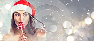 Christmas woman. Beauty model girl in Santa Claus hat with red lips and xmas lollipop candy. Closeup portrait