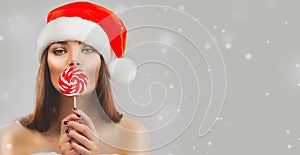 Christmas woman. Beauty model girl in Santa Claus hat with red xmas lollipop candy, gift. Sales. Surprised expression