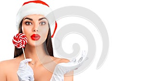 Christmas woman. Joyful model girl in Santa`s hat with lollipop candy pointing hand, proposing product. Surprised expression