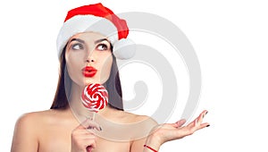 Christmas woman. Joyful model girl in Santa`s hat with lollipop candy pointing hand, proposing product. Surprised expression