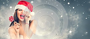 Christmas woman. Joyful model girl in Santa`s hat with red lips and lollipop candy in her hand