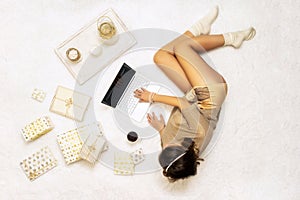 Christmas. Woman in sweater using laptop for searching gift ideas lying on the white carpet among the many wrapped boxes in white