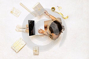 Christmas. Woman in sweater using laptop for searching gift ideas sitting on the white carpet among the many wrapped boxes