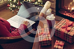 Christmas. Woman using laptop for searching gift ideas sitting at table with ready gift boxes and cup of cocoa and marshmallows