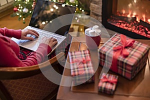 Christmas. Woman using laptop for searching gift ideas sitting at table with ready gift boxes and cup of cocoa and marshmallows