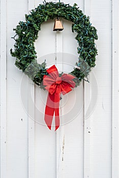 Christmas wreath made of artificial evergreen boughs and a light string, with red bow and cow bell, on a white wall