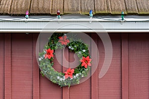 Christmas wreath made of artificial evergreen boughs and a light string, with red flowers and white snowflakes, on a red barn wall