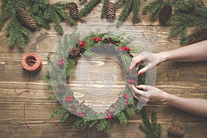 Christmas wreath made of spruce branches with holly berries on wooden board. Female hand decorated wreath. Top view.