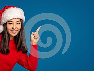 Christmas, xmas concept. Happy woman in santa claus hat pointing finger at copy space on blue background