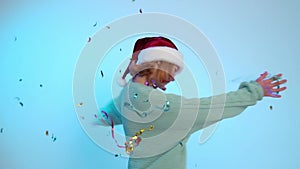 Christmas. Young girl dancing in the santa claus cap with confetti on the blue background.