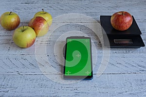 Chroma key green screen phone lies on a wooden table next to ripe apples, which are weighed on a kitchen scale.