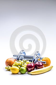 Chrome dumbbells surrounded with healthy fruits measuring tape on a white background with shadows.