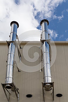 Chrome ventilation pipes on the outer wall of a beige industrial building against a blue sky background.
