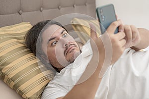 A chubby man in his 30s is shocked by a controversial social media post on his cellphone while lying on the bed photo