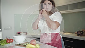 Chubby woman talking on the phone in kitchen as cooking fresh healthful salad. Portrait of positive young plus-size