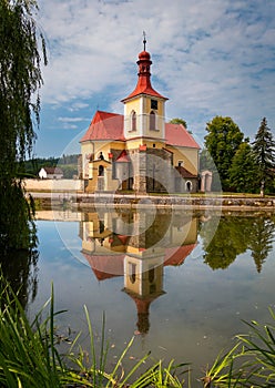 A church on the bank of a pond illuminated by the sun at sunset in the village of Mlade Briste, Czech