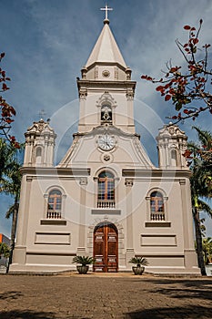 Church facade and belfry in front of a small cobblestone square with evergreen garden, in a sunny day at SÃ£o Manuel.