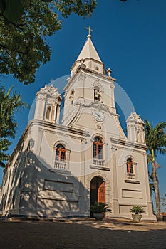 Church facade and belfry in front of a small cobblestone square with evergreen garden, in a sunny day at SÃ£o Manuel.