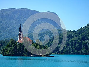 A church on a wooded island in a turquoise lake, with mountains behind