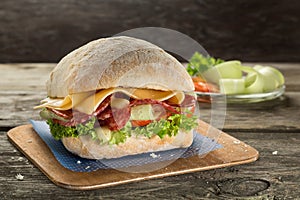 Ciabatta sandwich with salami, cheese, tomato and green salad close up on the wooden background