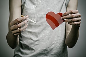 Cigarettes, addiction and public health topic: smoker holds the cigarette in his hand and a red heart on a dark background in the