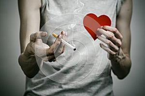 Cigarettes, addiction and public health topic: smoker holds the cigarette in his hand and a red heart on a dark background in the