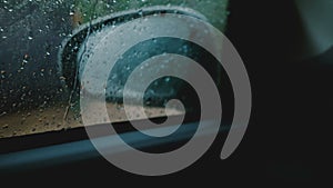 Cinematic close-up, defocused side view mirror is seen from inside the moving car, focus on rain drops on the window.