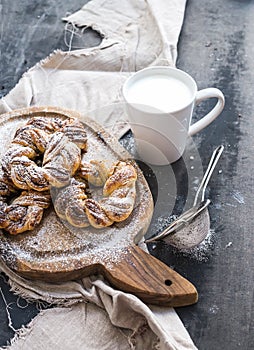 Cinnamon buns with sugar powder on rustic wooden board, mug of milk, dark grunge surface