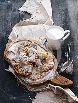 Cinnamon buns with sugar powder on rustic wooden board, mug of milk, dark grunge surface