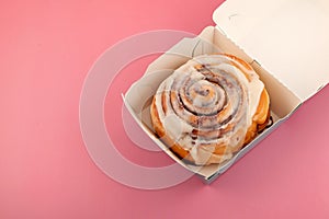 Cinnamon roll or cinnabon in paper box, top view. Homemade sweet traditional dessert buns with white cream sauce. Pink background