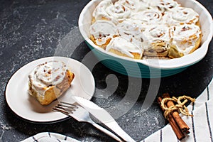 Cinnamon rolls or cinnabon, homemade recipe of sweet dessert buns with white cream sauce on dark stone background.