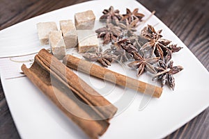 Cinnamon sticks, anise stars and pieces of brown sugar lie on a white square saucer