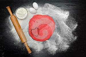 Circle of red dough with heart-shape cut-out. Black table sprinkled with flour, rolling pin and eggs